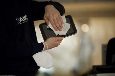A person is cleaning a tablet with a white cloth while holding a white face mask in the other hand. The individual is wearing a dark blue shirt. The background is blurred, suggesting an indoor setting.