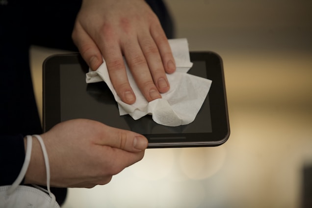 A person is cleaning a digital tablet with a white cloth, focusing on the screen. The hands are gently applying pressure to ensure the screen is wiped clean, possibly to remove fingerprints or dust.