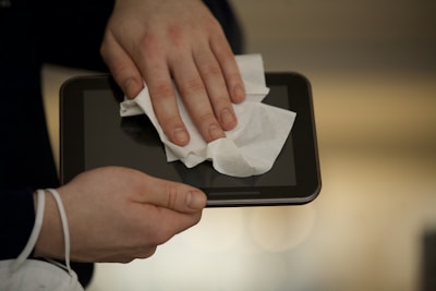 A person is cleaning a digital tablet with a white cloth, focusing on the screen. The hands are gently applying pressure to ensure the screen is wiped clean, possibly to remove fingerprints or dust.