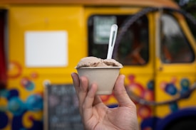 A hand holds a cup of ice cream with a spoon, set against the backdrop of a brightly colored yellow ice cream truck with vibrant, decorative patterns.