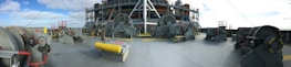 An industrial scene with large machinery and equipment placed on a ship deck. The machinery consists of several winches and other mechanical devices, accompanied by railings and safety barriers. The backdrop shows a blue sky with some scattered clouds.