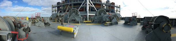 An industrial scene with large machinery and equipment placed on a ship deck. The machinery consists of several winches and other mechanical devices, accompanied by railings and safety barriers. The backdrop shows a blue sky with some scattered clouds.