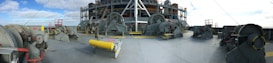 An industrial scene with large machinery and equipment placed on a ship deck. The machinery consists of several winches and other mechanical devices, accompanied by railings and safety barriers. The backdrop shows a blue sky with some scattered clouds.