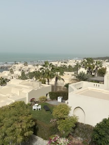 The image features a residential area with white, flat-roofed buildings surrounded by lush greenery and palm trees. In the foreground, a small garden with two white chairs and a sun lounger are visible. The background includes a glimpse of the distant, calm sea under a hazy sky.