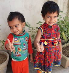 Two young children are standing side by side, smiling and making peace signs with their hands. They are wearing colorful clothing; one is in a red dress with floral patterns, and the other is in a green and red outfit with cartoon prints. Behind them are potted plants against a plain wall.
