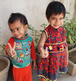 Two young children are standing side by side, smiling and making peace signs with their hands. They are wearing colorful clothing; one is in a red dress with floral patterns, and the other is in a green and red outfit with cartoon prints. Behind them are potted plants against a plain wall.