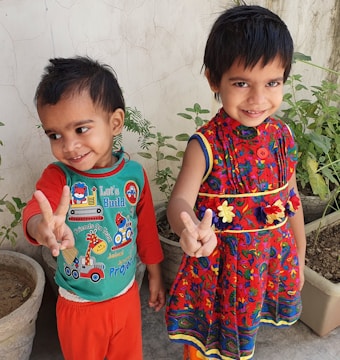 Two young children are standing side by side, smiling and making peace signs with their hands. They are wearing colorful clothing; one is in a red dress with floral patterns, and the other is in a green and red outfit with cartoon prints. Behind them are potted plants against a plain wall.