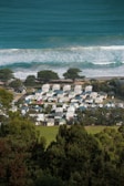 A coastal settlement with numerous houses or cabins closely packed together. The ocean is visible in the background with waves crashing onto the shore. Trees are scattered among the buildings, and there is lush green foliage in the foreground.