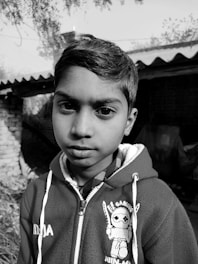A black-and-white portrait of a young person wearing a hoodie with a cartoon character design. The background features a rustic setting with brick and foliage.