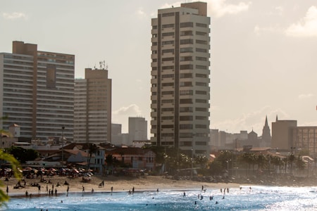 A city beach scene with tall modern buildings and a skyline in the background. People are enjoying activities in the ocean and on the sandy shore in the foreground, while umbrellas dot the beach area.