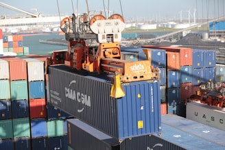 A cargo container lifting mechanism is hoisting a blue shipping container with 'CMA CGM' written on it. The area is surrounded by stacks of colorful shipping containers in various sizes. In the background, there is a harbor with cranes and wind turbines visible, along with some industrial buildings.