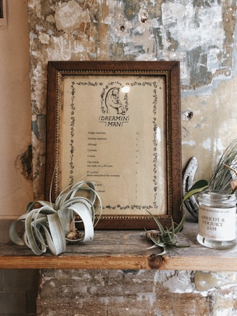 A rustic interior setting featuring a framed menu titled 'Dreamini Man', placed against a textured, peeling wall. The menu lists various coffee options. On the shelf, there's a glass jar labeled 'Apricot & Verjuice Jam' and some decorative green plants with a light grayish hue, possibly air plants.