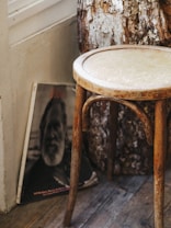 A rustic wooden stool is placed on a wooden floor near a textured tree stump and a black-and-white portrait of an older man leaning against the wall. The scene has a natural and vintage aesthetic with warm tones.