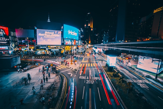 A vibrant urban scene at night featuring a bustling intersection with brightly lit billboards and advertisements. The streets are busy with blurred lights from passing vehicles, giving a sense of motion. People are seen walking on the sidewalks, and there are visible signs for popular brands such as McDonald's and Starbucks.