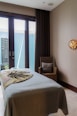 A tranquil massage room with a neatly made massage table covered in a neutral-toned sheet and a decorative blanket. A wicker chair with a cushion is placed beside the table. A serene Buddha statue is visible outside the glass doors that allow natural light to enter. The room features a calming ambiance with minimalist decor and a wall light adding warmth to the space.