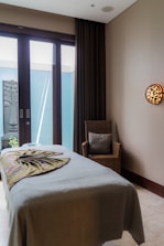 A tranquil massage room with a neatly made massage table covered in a neutral-toned sheet and a decorative blanket. A wicker chair with a cushion is placed beside the table. A serene Buddha statue is visible outside the glass doors that allow natural light to enter. The room features a calming ambiance with minimalist decor and a wall light adding warmth to the space.