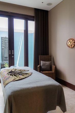 A tranquil massage room with a neatly made massage table covered in a neutral-toned sheet and a decorative blanket. A wicker chair with a cushion is placed beside the table. A serene Buddha statue is visible outside the glass doors that allow natural light to enter. The room features a calming ambiance with minimalist decor and a wall light adding warmth to the space.