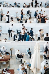 A gathering of people, mostly men, wearing traditional religious garments and masks are engaged in prayer and reading, in an outdoor setting with white plastic chairs and tables. Some individuals are seated, while others are standing, showing diverse activities including reading from religious texts and personal reflection.