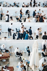 A gathering of people, mostly men, wearing traditional religious garments and masks are engaged in prayer and reading, in an outdoor setting with white plastic chairs and tables. Some individuals are seated, while others are standing, showing diverse activities including reading from religious texts and personal reflection.
