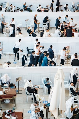 A gathering of people, mostly men, wearing traditional religious garments and masks are engaged in prayer and reading, in an outdoor setting with white plastic chairs and tables. Some individuals are seated, while others are standing, showing diverse activities including reading from religious texts and personal reflection.
