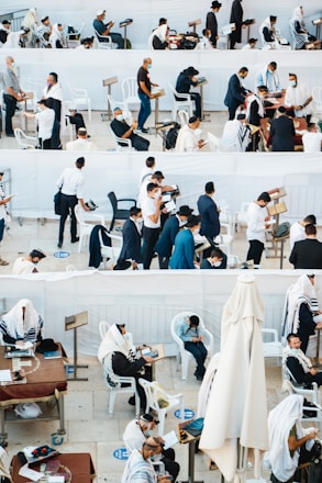 A gathering of people, mostly men, wearing traditional religious garments and masks are engaged in prayer and reading, in an outdoor setting with white plastic chairs and tables. Some individuals are seated, while others are standing, showing diverse activities including reading from religious texts and personal reflection.