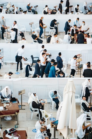 A gathering of people, mostly men, wearing traditional religious garments and masks are engaged in prayer and reading, in an outdoor setting with white plastic chairs and tables. Some individuals are seated, while others are standing, showing diverse activities including reading from religious texts and personal reflection.