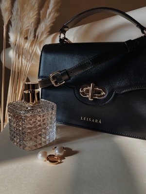 A black leather handbag with a gold clasp sits next to a geometric-patterned glass perfume bottle. Two pearl earrings with gold accents are placed on a light surface, while soft, dried pampas grass in the background adds a natural element.