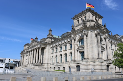 A grand, historic building featuring classical architecture with large columns and intricate details. The structure is topped with German flags, suggesting its national significance. The setting is under a clear blue sky, with barriers visible in the foreground, possibly for security or crowd control.
