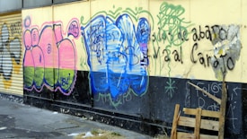 Colorful graffiti is painted on a wall featuring abstract shapes and letters in vibrant pink, green, and blue hues. The wall is partially worn and cracked, with wooden pallets leaning against it. Black spray paint adds more text and symbols, contributing to a gritty urban feel.