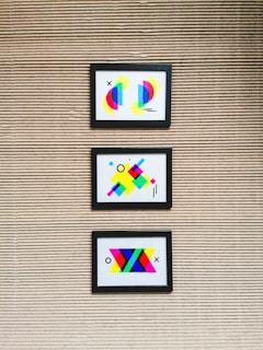 Three framed abstract artworks featuring bright geometric shapes are mounted on a textured wall with horizontal lines. Each artwork is unique, comprising colorful elements such as circles, rectangles, and lines in various vibrant colors like neon pink, blue, yellow, and green.