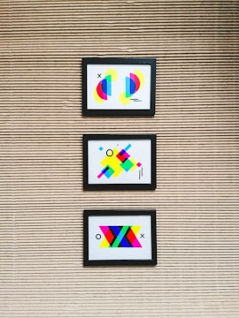 Three framed abstract artworks featuring bright geometric shapes are mounted on a textured wall with horizontal lines. Each artwork is unique, comprising colorful elements such as circles, rectangles, and lines in various vibrant colors like neon pink, blue, yellow, and green.