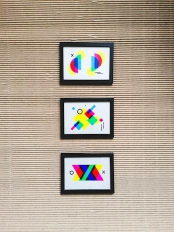 Three framed abstract artworks featuring bright geometric shapes are mounted on a textured wall with horizontal lines. Each artwork is unique, comprising colorful elements such as circles, rectangles, and lines in various vibrant colors like neon pink, blue, yellow, and green.