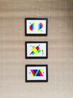 Three framed abstract artworks featuring bright geometric shapes are mounted on a textured wall with horizontal lines. Each artwork is unique, comprising colorful elements such as circles, rectangles, and lines in various vibrant colors like neon pink, blue, yellow, and green.