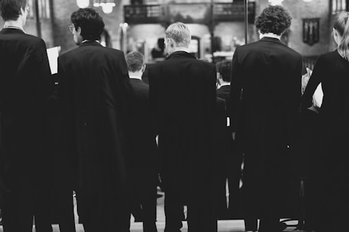 A group of people dressed in formal attire, possibly choir members, stand in rows with their backs to the camera in a dimly lit room. The setting appears to be a church or concert hall with ornate lighting and a decorative balcony visible in the background.
