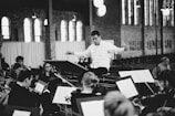 A conductor in a white shirt is leading an orchestra of musicians playing string instruments in a spacious hall with large windows. The musicians are focused on their sheet music, creating an atmosphere of concentration and harmony.