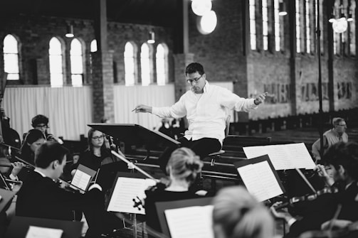 A conductor in a white shirt is leading an orchestra of musicians playing string instruments in a spacious hall with large windows. The musicians are focused on their sheet music, creating an atmosphere of concentration and harmony.