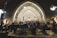 An orchestra performs in a historic arched hall with musicians dressed in formal attire. The conductor stands at the center, leading the ensemble. The backdrop features a stone wall with illuminated arches, adding a serene and grand atmosphere to the setting.