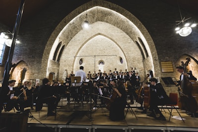 An orchestra performs in a historic arched hall with musicians dressed in formal attire. The conductor stands at the center, leading the ensemble. The backdrop features a stone wall with illuminated arches, adding a serene and grand atmosphere to the setting.