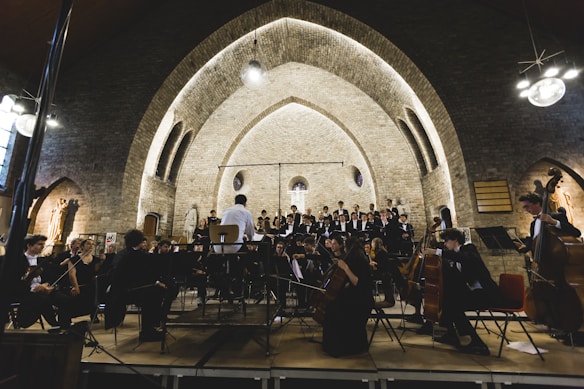 An orchestra performs in a historic arched hall with musicians dressed in formal attire. The conductor stands at the center, leading the ensemble. The backdrop features a stone wall with illuminated arches, adding a serene and grand atmosphere to the setting.