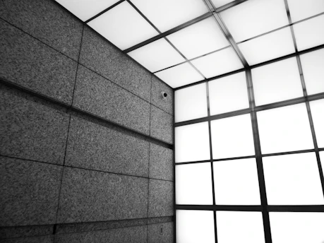 An architectural interior featuring a modern, industrial design with large stone or concrete panels on the walls. The ceiling consists of a grid of square lights, casting a diffused glow.