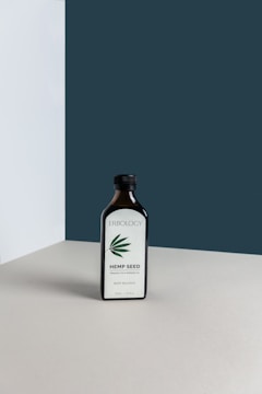 A dark glass bottle labeled 'Erbology Hemp Seed' is placed on a neutral-colored surface with a minimalist backdrop. The bottle has a simple design with a green hemp leaf illustration on the label.