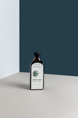 A dark glass bottle labeled 'Erbology Hemp Seed' is placed on a neutral-colored surface with a minimalist backdrop. The bottle has a simple design with a green hemp leaf illustration on the label.