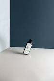 A bottle labeled 'ERBOLOGY Hemp Seed Oil' is leaning at an angle on a minimalist, neutral-toned surface. The background features walls with a mix of dark blue and light grey colors, creating a stark contrast.