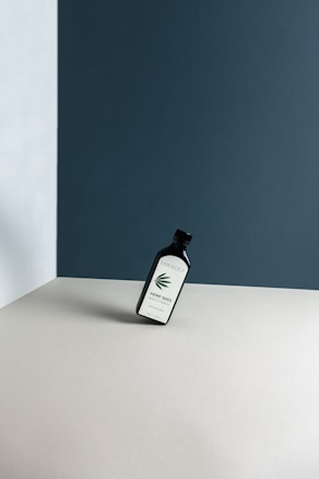 A bottle labeled 'ERBOLOGY Hemp Seed Oil' is leaning at an angle on a minimalist, neutral-toned surface. The background features walls with a mix of dark blue and light grey colors, creating a stark contrast.