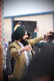 A person wearing a green turban and a light brown suit appears to be dancing or celebrating with arms raised. The atmosphere is lively, with other individuals in the background also appearing engaged, possibly at an event or gathering.