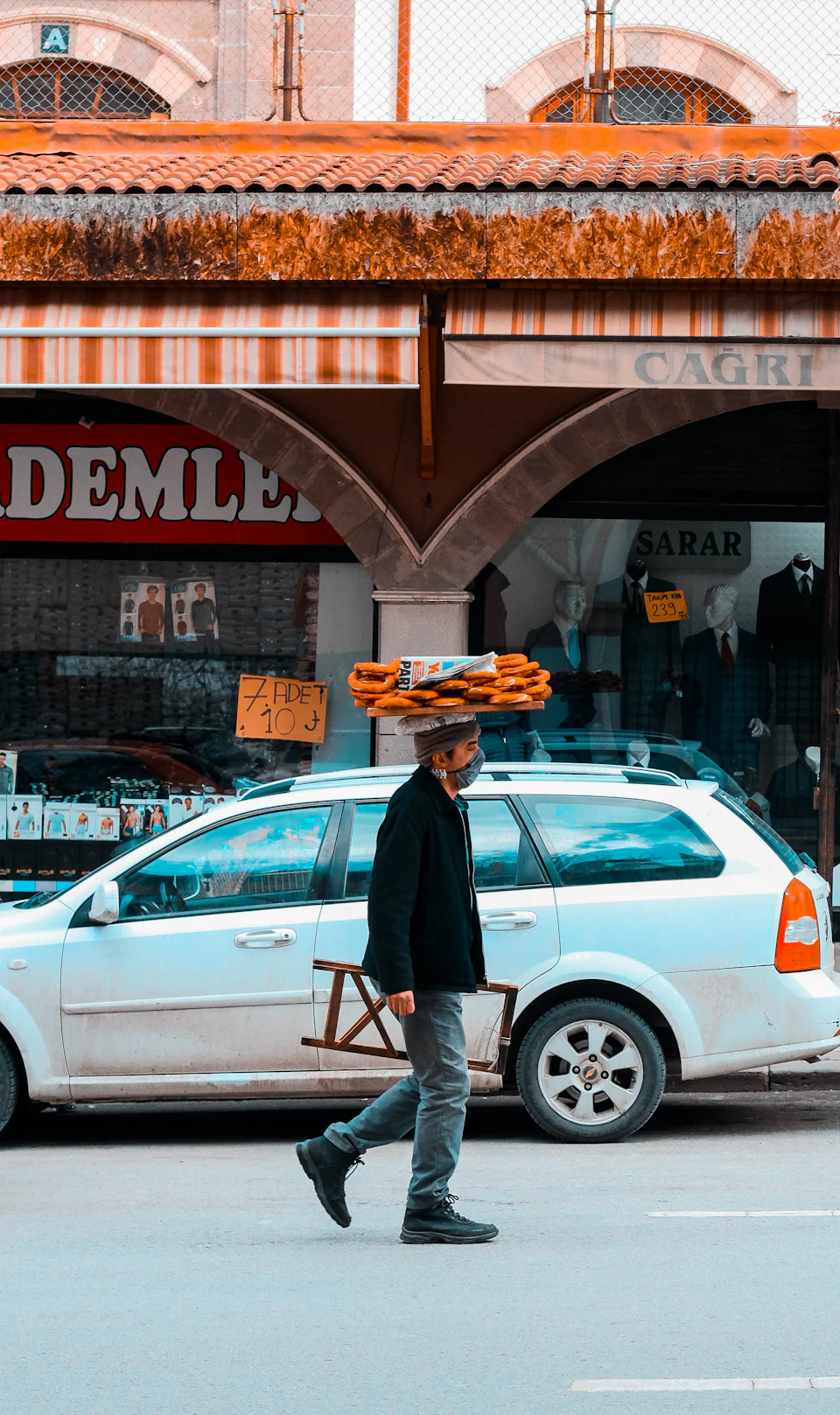 The image depicts a man walking down a street, carrying a large bundle on his head and balancing it with one hand while holding a wooden crate in the other. The man is dressed in a black jacket and gray pants, and he appears to be moving towards a white car parked nearby. In the background, there are buildings lining the street, including a store named "DEMELLE" that has a red awning over its entrance.
