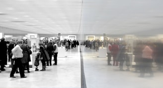 A large group of people gathered in an art gallery or museum, observing various displays and artworks. The space is expansive and well-lit, with a reflective wall dividing the scene, creating a symmetrical effect.