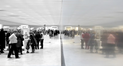 A large group of people gathered in an art gallery or museum, observing various displays and artworks. The space is expansive and well-lit, with a reflective wall dividing the scene, creating a symmetrical effect.
