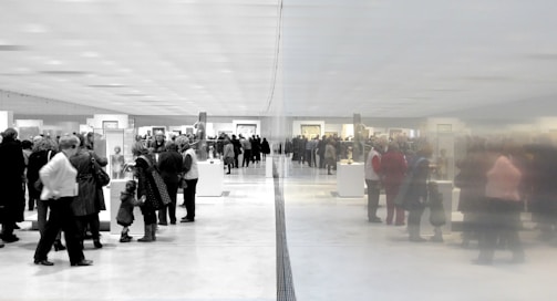 A large group of people gathered in an art gallery or museum, observing various displays and artworks. The space is expansive and well-lit, with a reflective wall dividing the scene, creating a symmetrical effect.