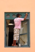 A person wearing a pink shirt and plaid shorts is balancing on the edge of an open window while painting the upper part of the frame. The window has a green frame with multiple small circular panes at the top. There is a paint can and brush being used for painting. The curtains inside the room are drawn, and the overall wall color is a peach or light orange.